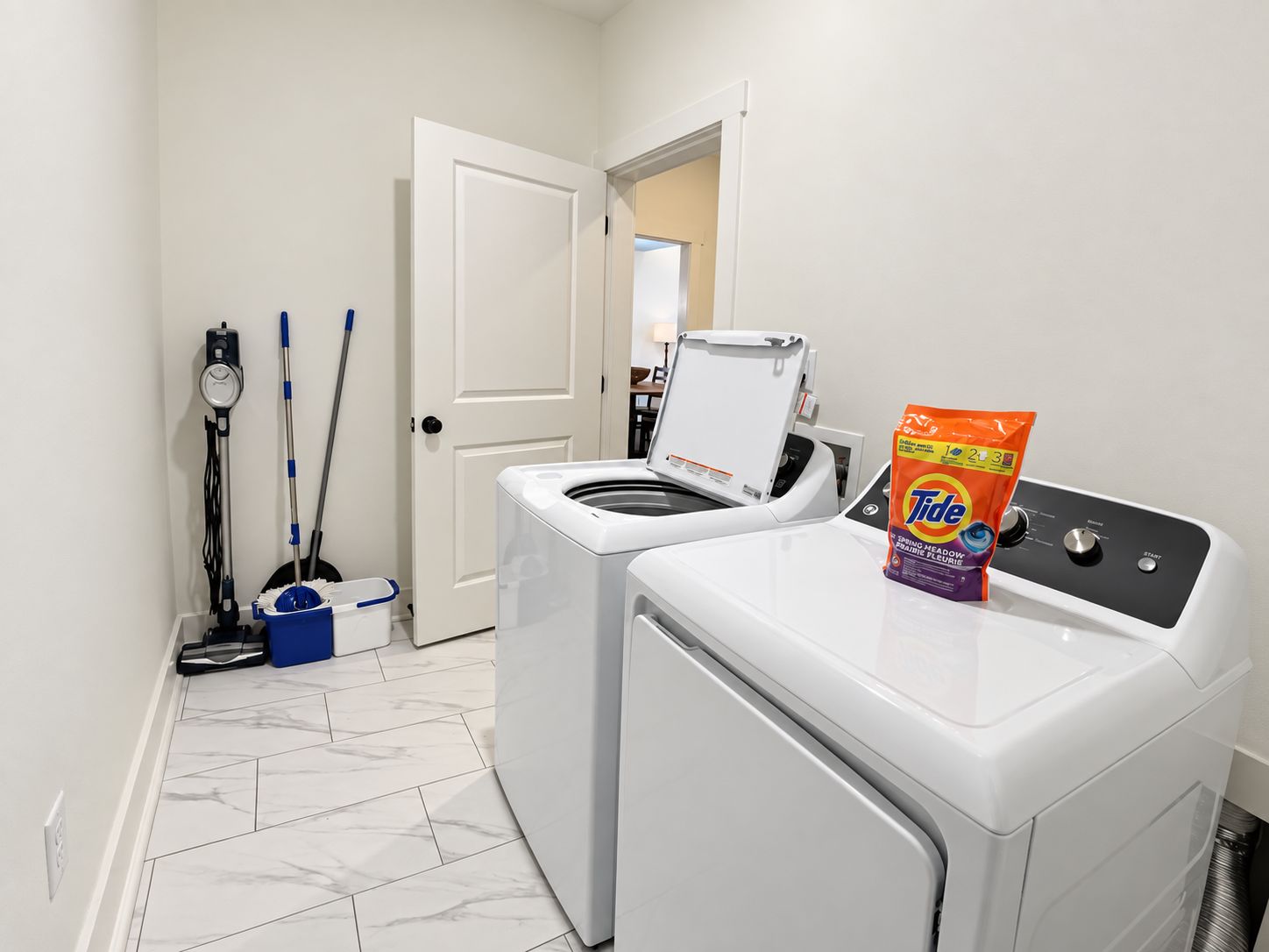 A laundry room with a front-loading washer and dryer, a mop, a bucket, and a bag of Tide detergent.