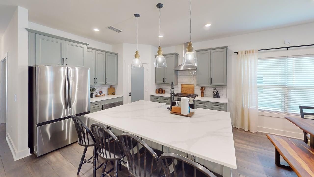 Une cuisine moderne dotée d’un grand îlot central, d’appareils électroménagers en acier inoxydable, de luminaires suspendus et offrant une vue sur l’extérieur.