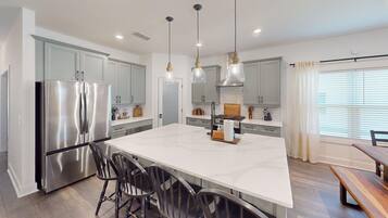 Une cuisine moderne dotée d’un grand îlot central, d’appareils électroménagers en acier inoxydable, de luminaires suspendus et offrant une vue sur l’extérieur.