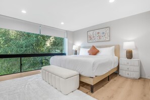 A modern bedroom with a large window, a bed with white bedding, a bench, and a bedside table with a lamp. - Mulholland Ridge By Alora Reserve (Los Angeles)