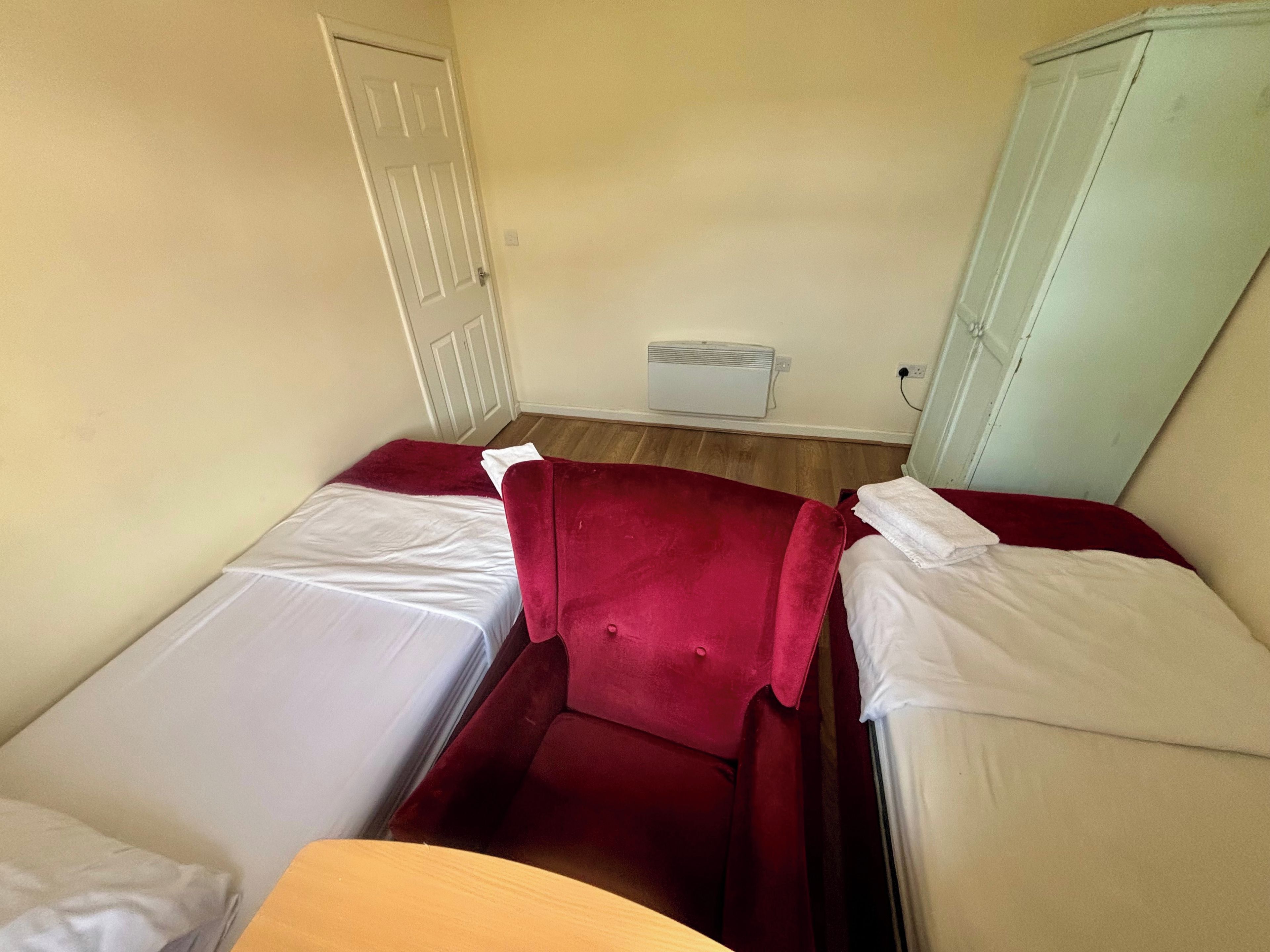 A small, single-bed room with a red headboard, a white bedside table, and a white wall-mounted heater.