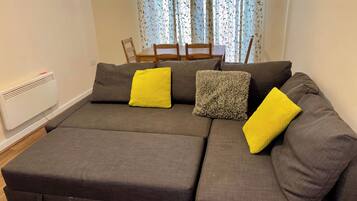 A sectional sofa with two yellow cushions and a textured grey pillow, a white radiator, and a dining area with a table and chairs.