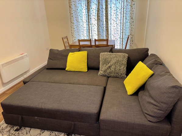A sectional sofa with two yellow cushions and a textured grey pillow, a white radiator, and a dining area with a table and chairs. - Apartment Sleeps 5 With Wifi (Wolverhampton)