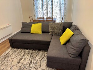 A sectional sofa with two yellow pillows and a grey one, a patterned rug, and a dining area with a table and chairs in the background. - Apartment Sleeps 5 With Wifi (Wolverhampton)
