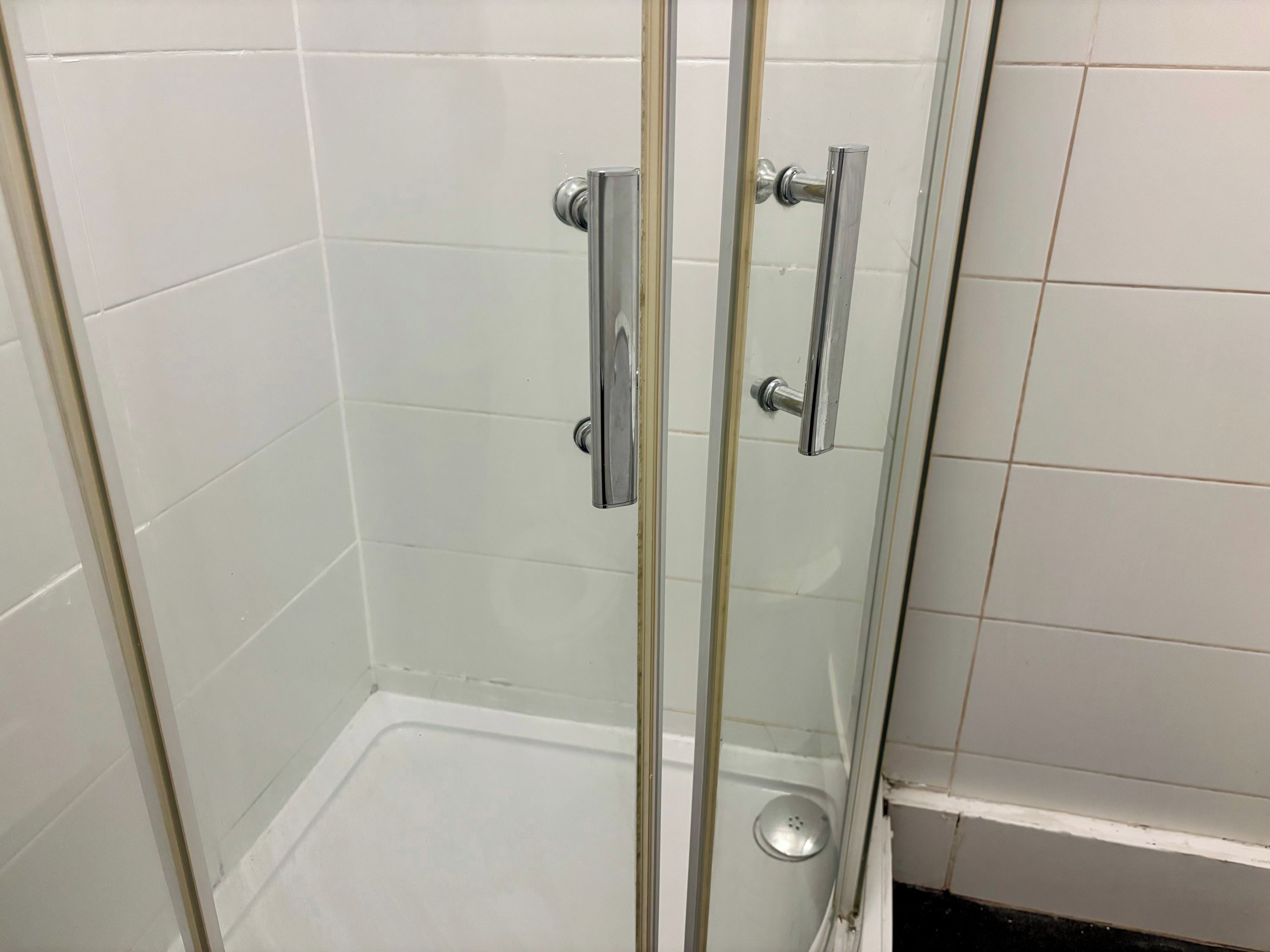 A glass-enclosed shower stall with a white tiled wall and a metal handle.