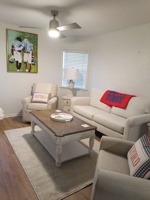 A living room with a ceiling fan, a window with blinds, a sofa with a red throw blanket, and a coffee table with a magazine. - Updated 1-bedroom condo, less than a mile from Stadium, LG covered outdoor space (Oxford)