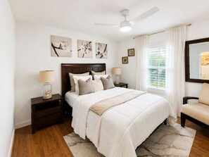 Ein Schlafzimmer mit einem Bett, Nachttischen, einem Deckenventilator, Kunstwerken an der Wand und einem Fenster mit Vorhängen.