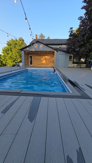 A swimming pool with a wooden deck and a building in the background. - Country Farmhouse Retreat with Pool (Uxbridge)