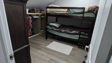A dormitory room with bunk beds, a wardrobe, a closet with hanging clothes, and a small desk.