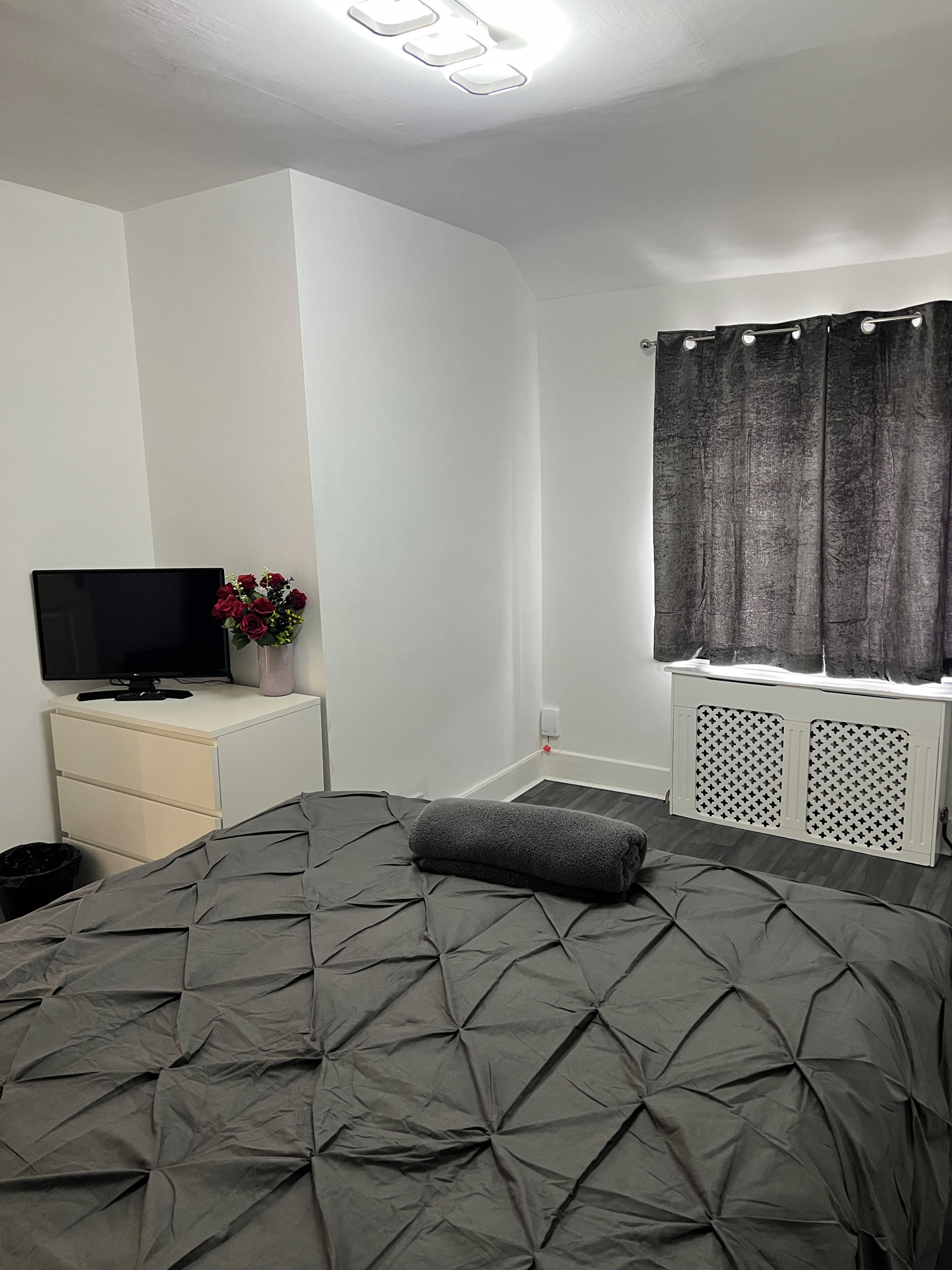A bedroom with a bed, a TV on a stand, a window with curtains, and a radiator.