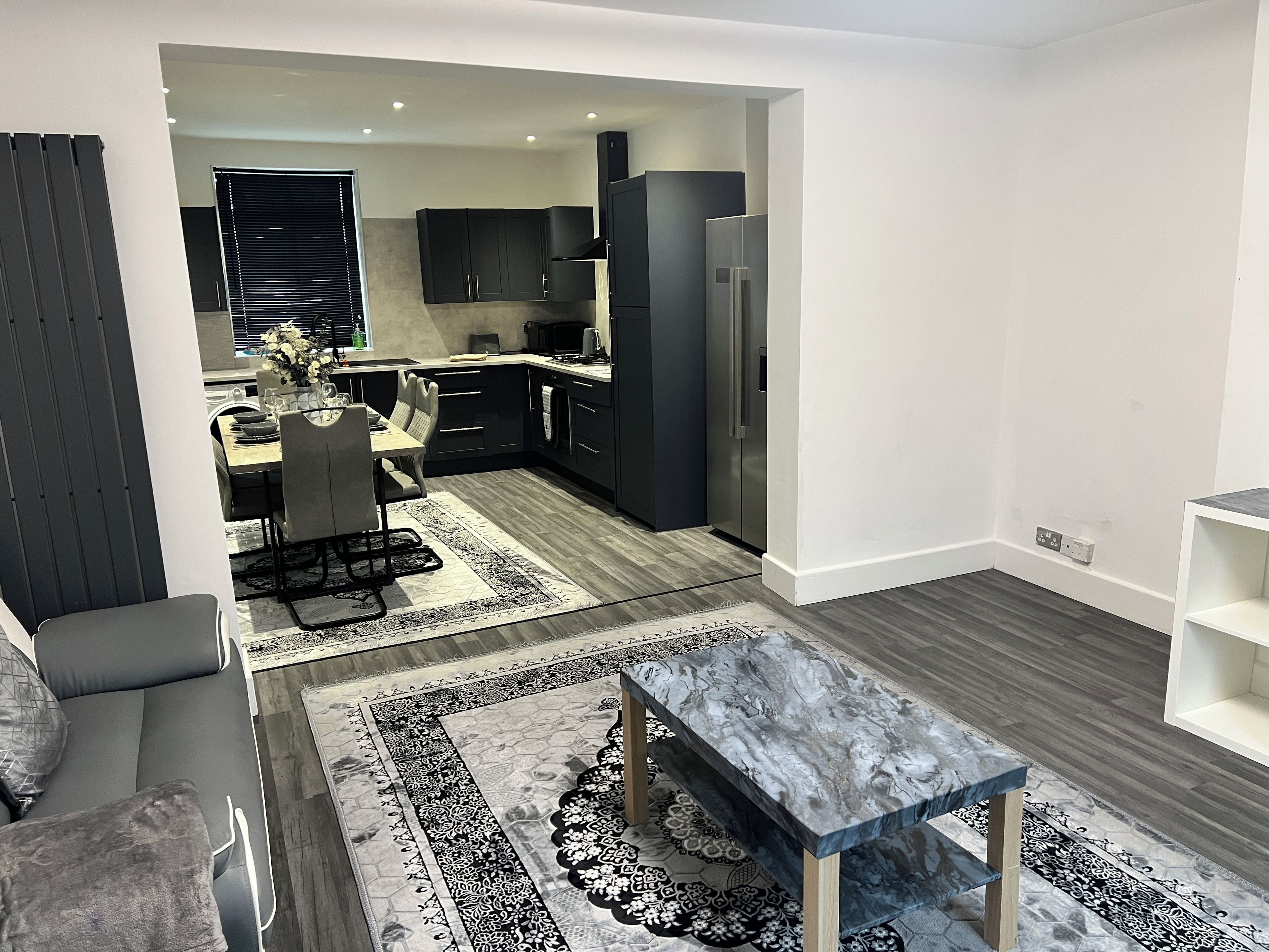 A modern living room with a marble coffee table, a dining area with a table and chairs, and a kitchen with dark cabinetry and appliances.