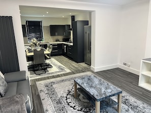 Uma sala de estar moderna com uma mesa de centro de mármore, um espaço de refeições com mesa e cadeiras e uma cozinha com armários e eletrodomésticos escuros.