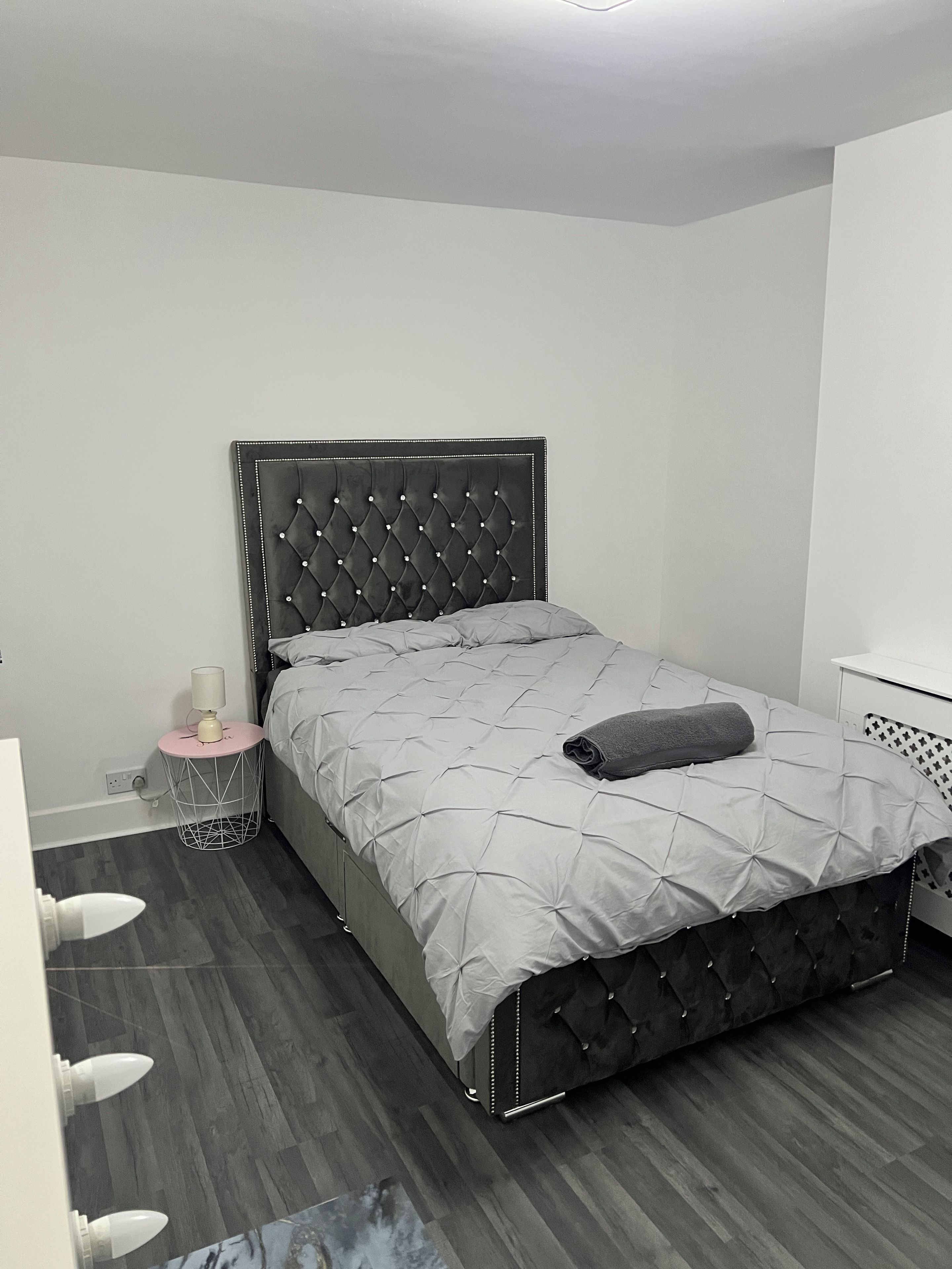 Un dormitorio con cabecera capitonada, colcha acolchada gris, mesita de noche con lámpara y una mesita auxiliar redonda.