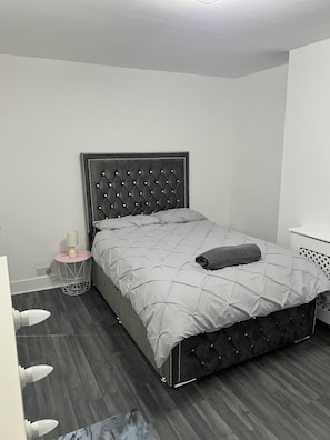 A bedroom with a tufted headboard, a grey quilted bedspread, a bedside table with a lamp, and a small round side table.