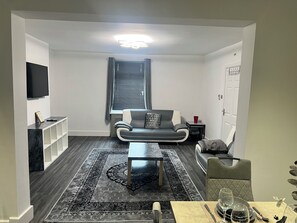 A modern living room with a sofa, a coffee table, a TV mounted on the wall, and a dining area with a table set for two.