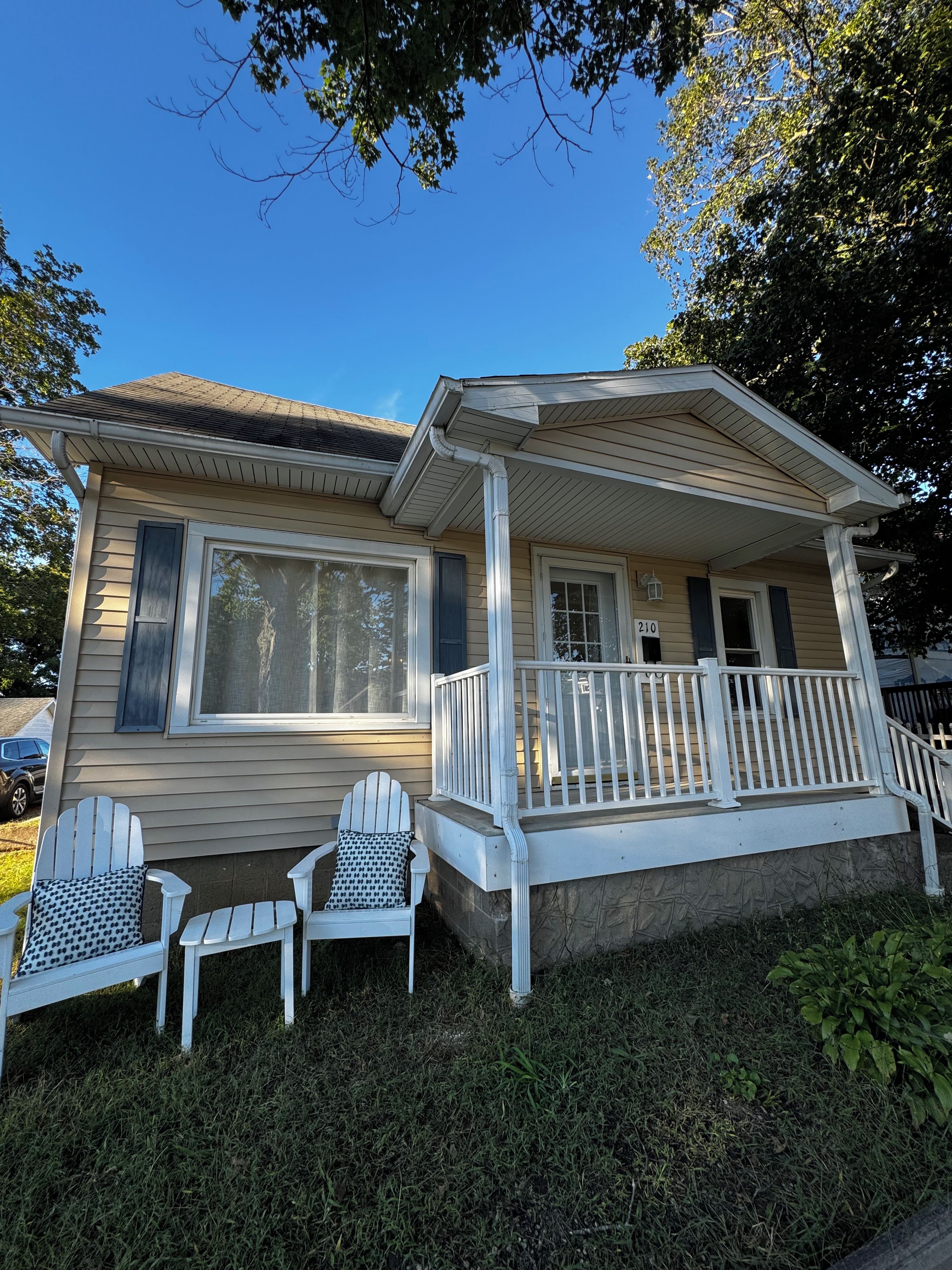 A single-story house with a front porch, two white Adirondack chairs, and a small front yard.