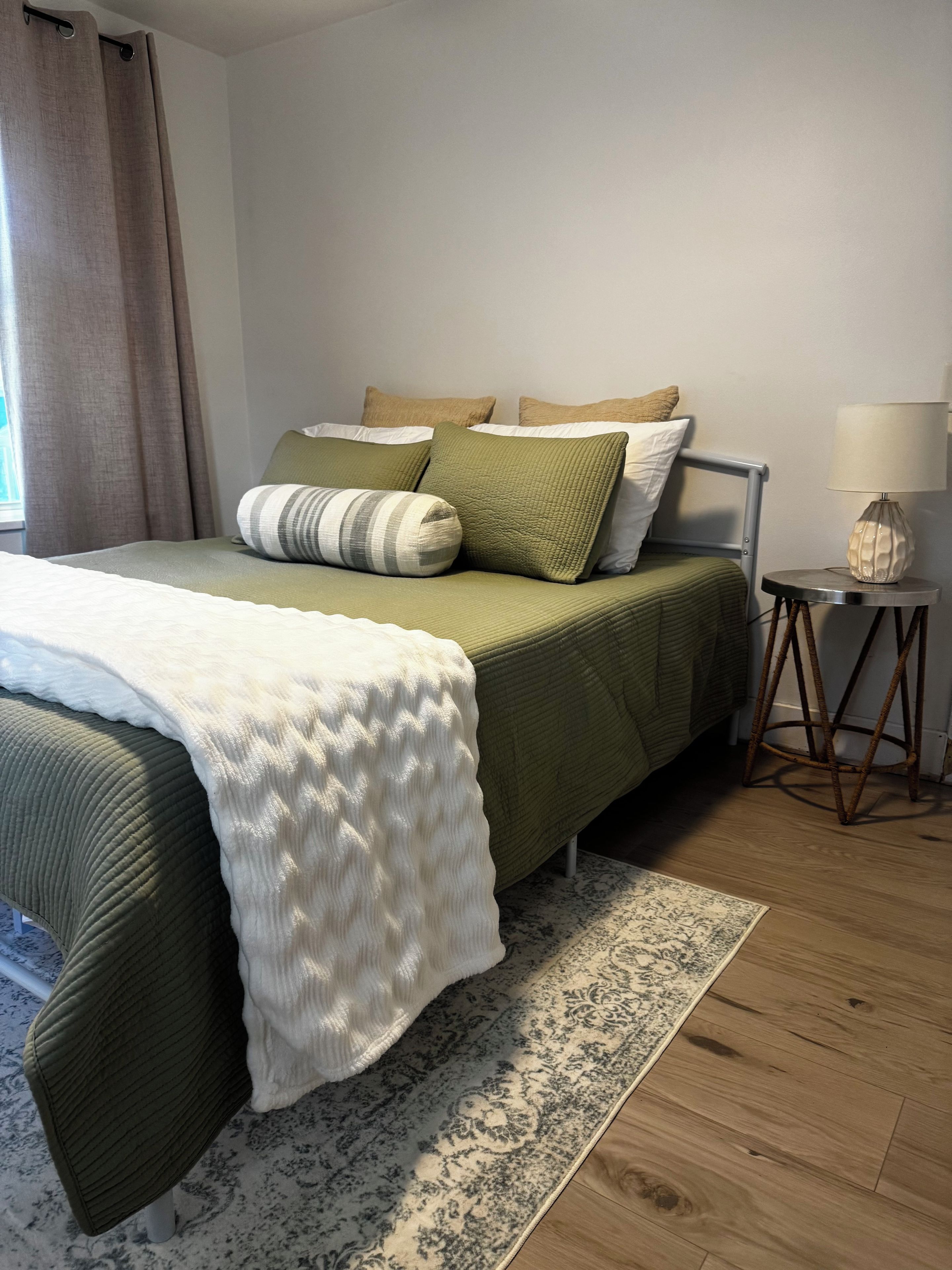 A neatly made bed with a white and green bedspread, a patterned rug, and a bedside lamp on a small wooden table.