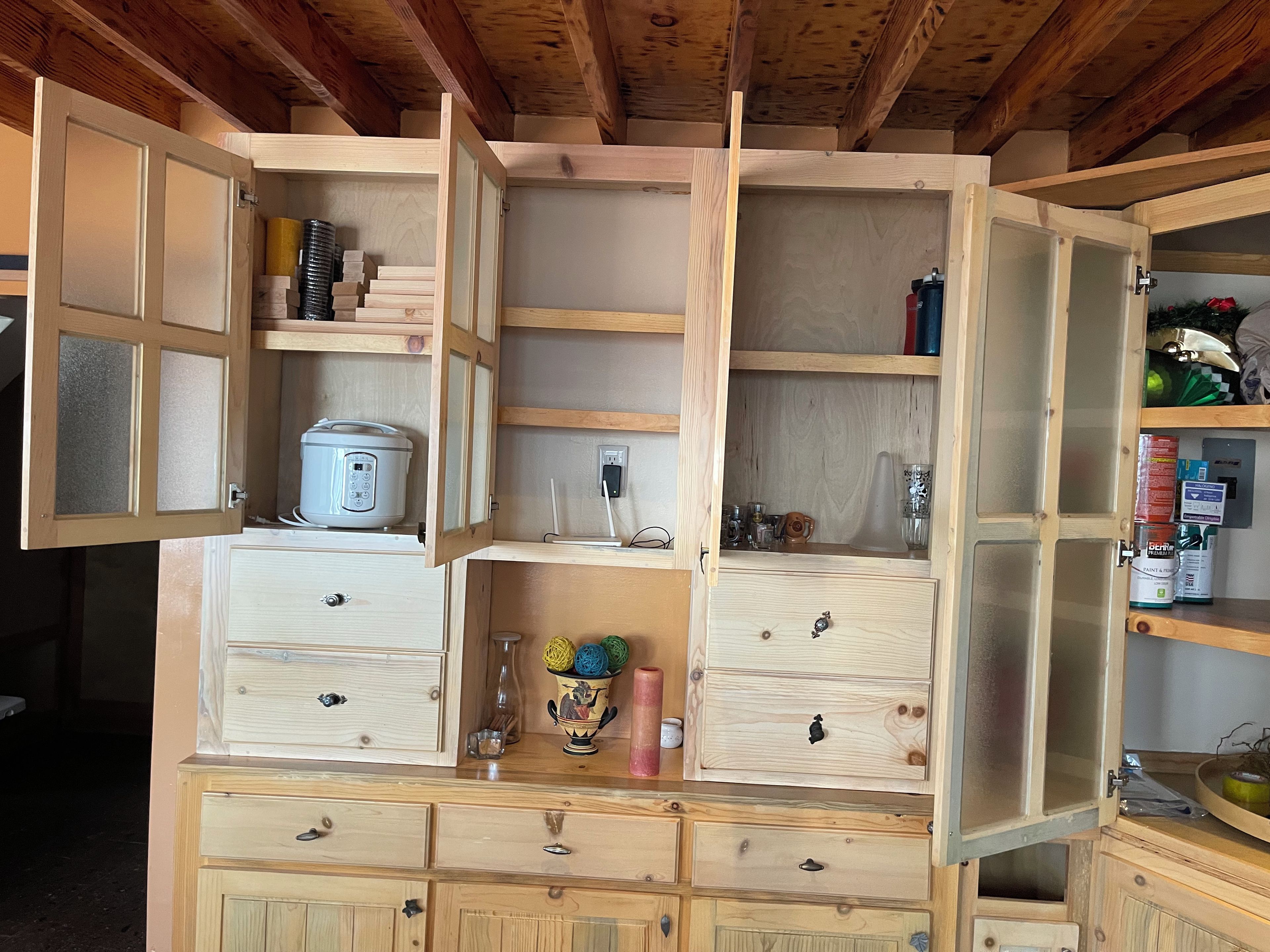 A wooden cabinet with multiple compartments, including a section with a white appliance, a section with glassware, and a section with various containers.