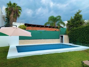 A swimming pool with a tiled edge, surrounded by grass, a palm tree, and a green fence. - Lomas Cocoyoc Private pool / barbecue / Wi-Fi / pet-friendly (Fraccionamiento Lomas de Cocoyoc)