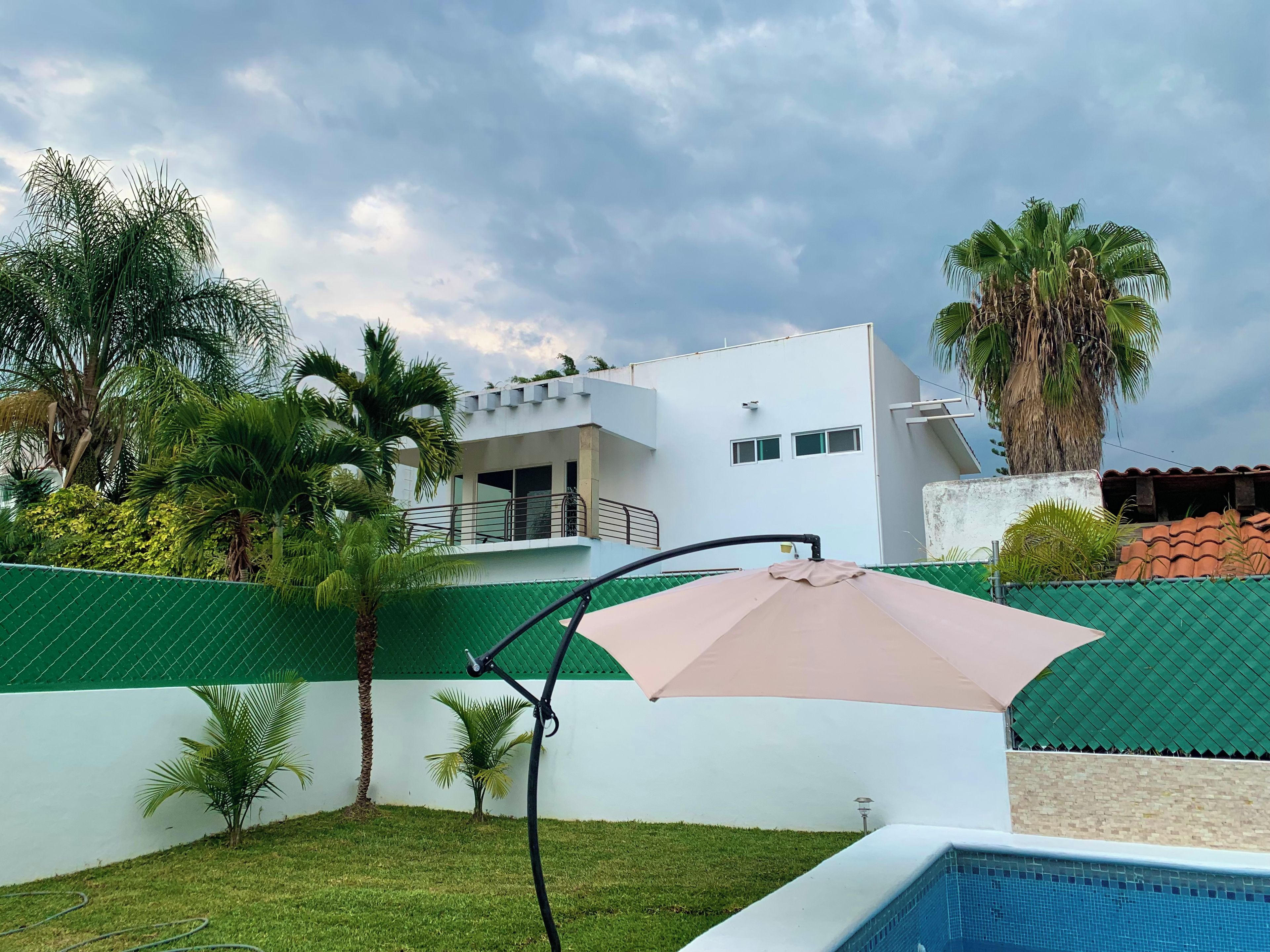 A modern building with a swimming pool, surrounded by greenery and a cloudy sky.