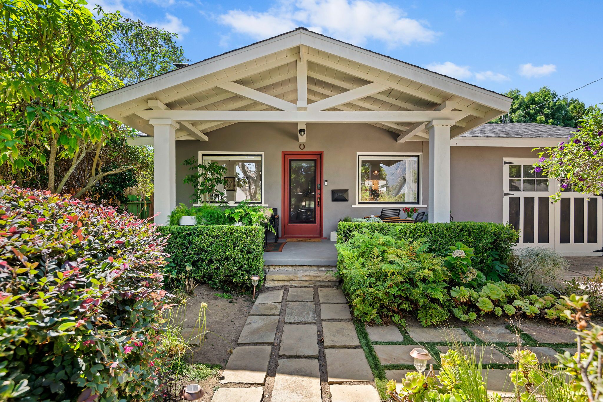 A house with a front porch, a red door, and a garden with various plants.