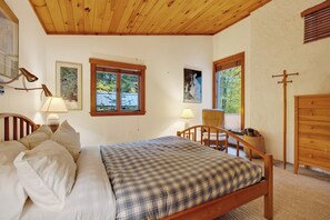 Ein Schlafzimmer mit einem hölzernen Bett, zwei Nachttischlampen, einer Holzkommode und einem Fenster mit Blick auf Bäume.