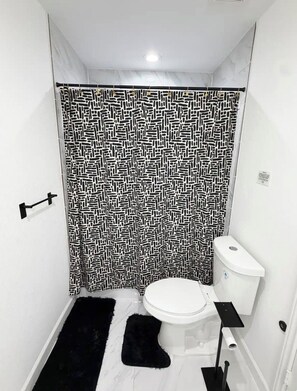 Een badkamer met een gordijn met een patroon, een toilet, een douchetoilet en een zwart vloerkleed.