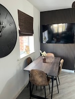 A dining area with a large wall clock, a flat-screen TV, and a window with a view of the outside.