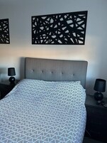 Un dormitorio con cabecera tapizada en gris, colcha estampada, dos mesitas de noche con lámparas y paneles decorativos en la pared.