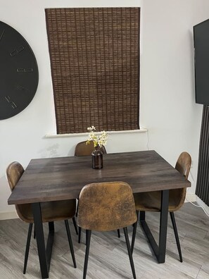 Un comedor con una mesa rectangular, cuatro sillas, un jarrón con flores, un reloj grande y un televisor en la pared.