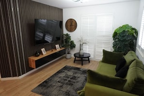 A living room with a flat-screen TV, a wooden entertainment unit, a green sofa, a round coffee table, and a large plant. - 2B 2B Haydock Flat M6 J23 A580 Gated Parking (St Helens)