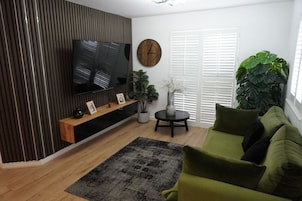 Una sala de estar con un televisor de pantalla plana, un mueble de entretenimiento de madera, un sofá verde, una mesa de centro redonda y una planta grande.