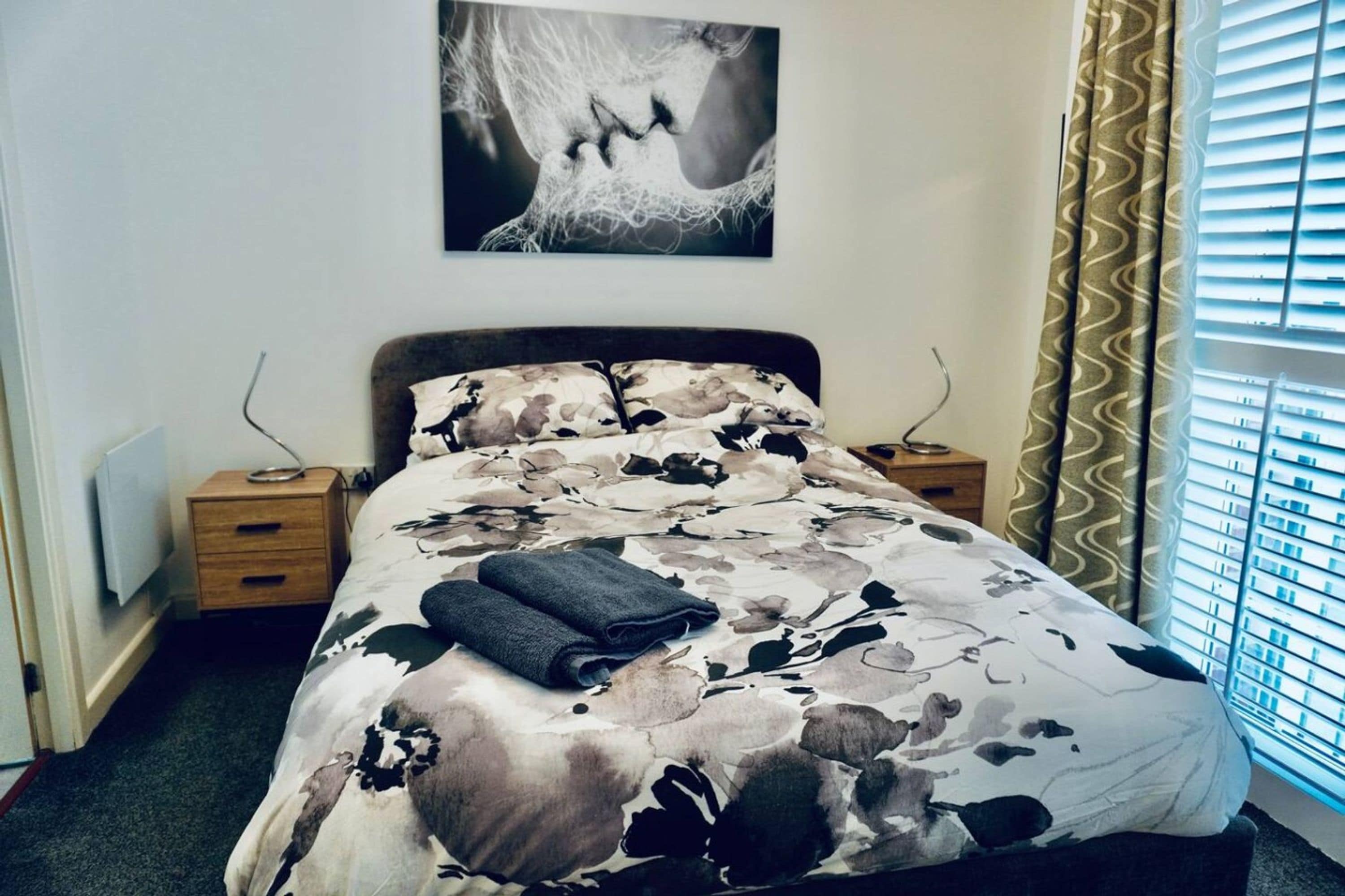 A bedroom with a bed, a nightstand, a wall-mounted artwork, and a window with curtains.
