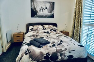 Un dormitorio con cama, mesita de noche, una obra de arte en la pared y una ventana con cortinas.