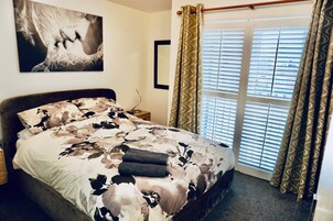 Un dormitorio con cama, mesita de noche, ventana con persianas y un cuadro colgado en la pared.