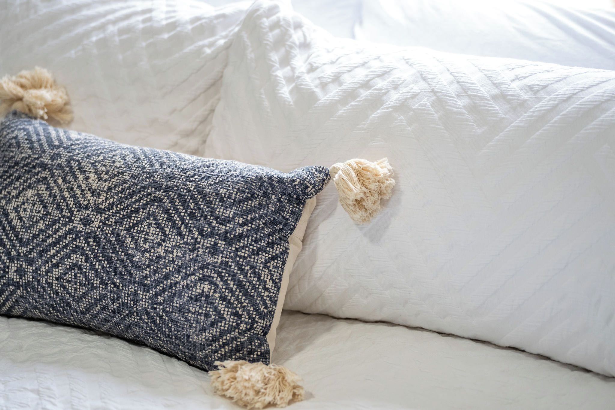 Een bed met witte lakens en een decoratief kussen met franjes.