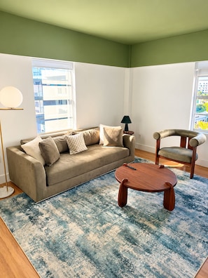 A living room with a sofa, a coffee table, and a chair. There is a lamp and a window with a view of buildings. - Deluxe King Suite, Beach Access & Prime Location (Miami Beach)