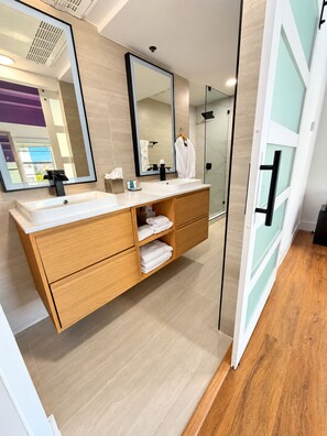 A modern bathroom with a large mirror, a white sink, wooden cabinets, and a glass shower enclosure. - Deluxe King Suite, Beach Access & Prime Location (Miami Beach)