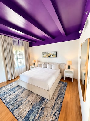 A bedroom with a large bed, two bedside tables with lamps, a ceiling with purple beams, and a window with curtains. - Deluxe King Suite, Beach Access & Prime Location (Miami Beach)