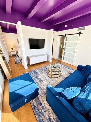 A modern living room with a blue sofa, a glass coffee table, and a large flat-screen TV mounted on the wall. - Deluxe King Suite, Beach Access & Prime Location (Miami Beach)