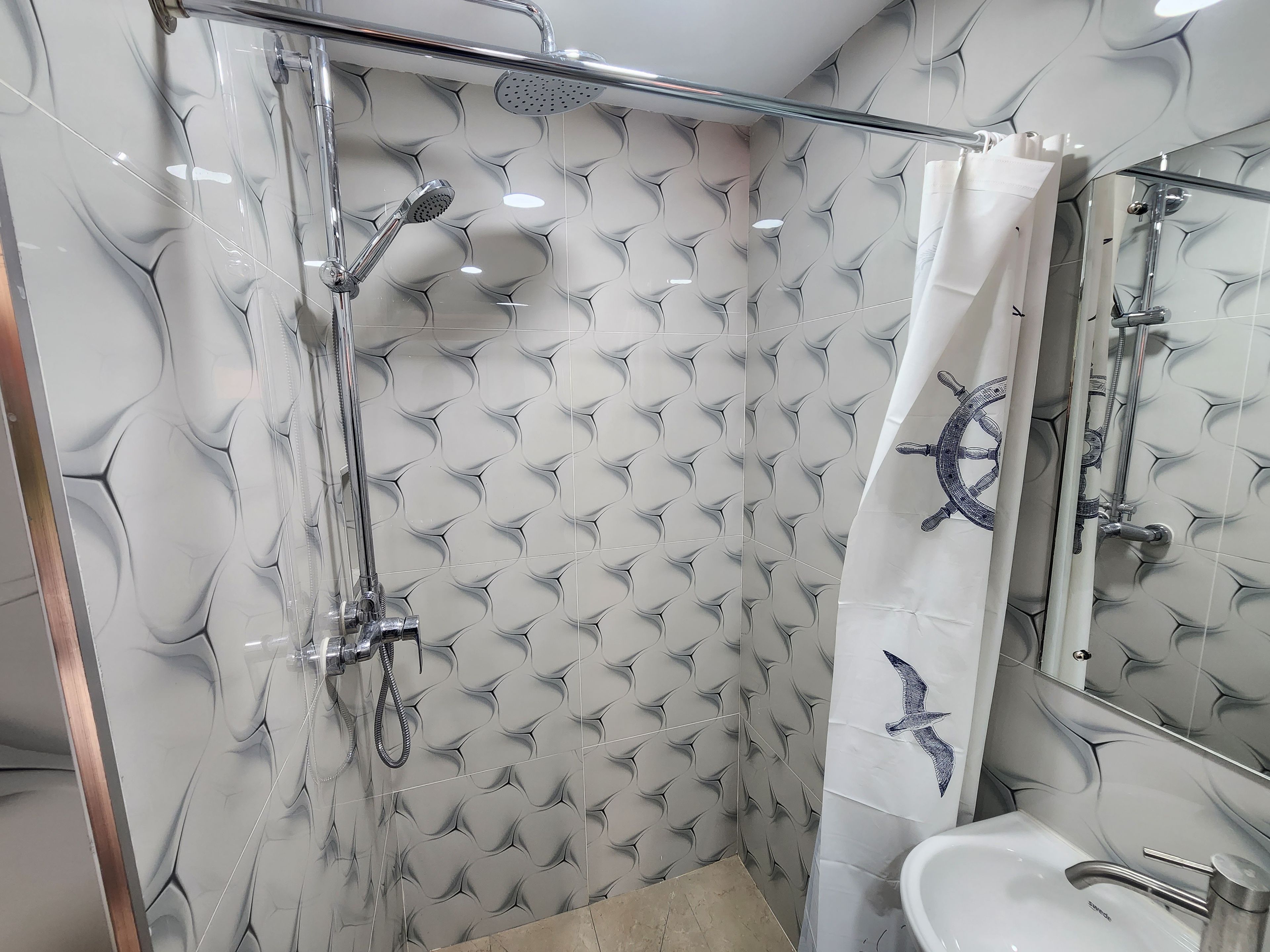 A bathroom with a glass-enclosed shower, a white sink, and a mirror with a ship wheel design.
