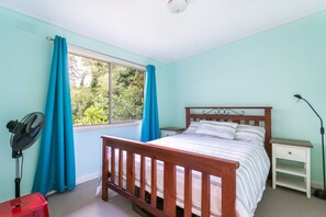 Een slaapkamer met een houten bed, een nachtkastje, een bedlamp, een ventilator en een raam met gordijnen.