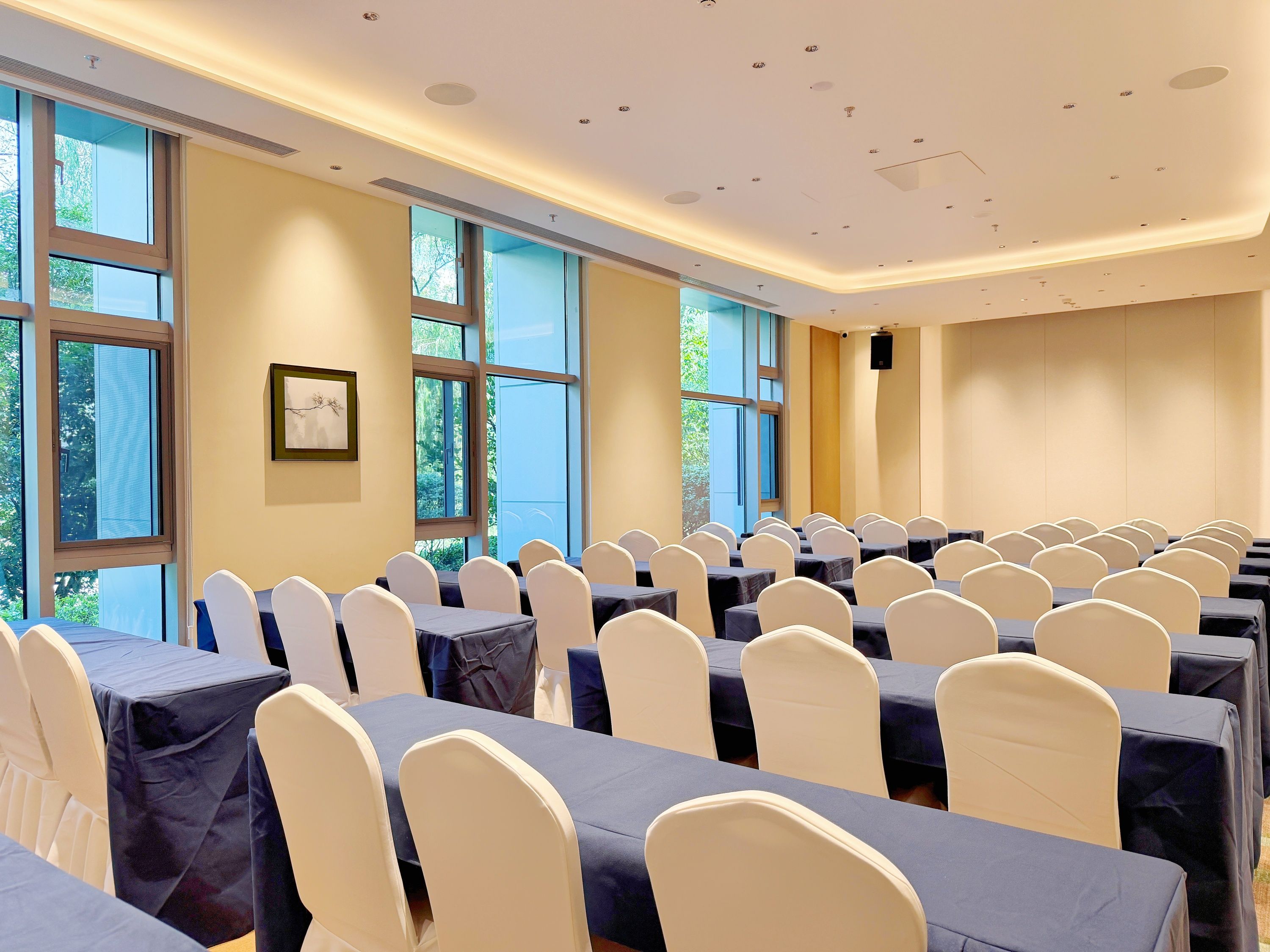 A conference room with rows of tables and chairs, large windows, and a speaker on the wall.