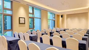 A conference room with rows of tables and chairs, large windows, and a speaker on the wall.