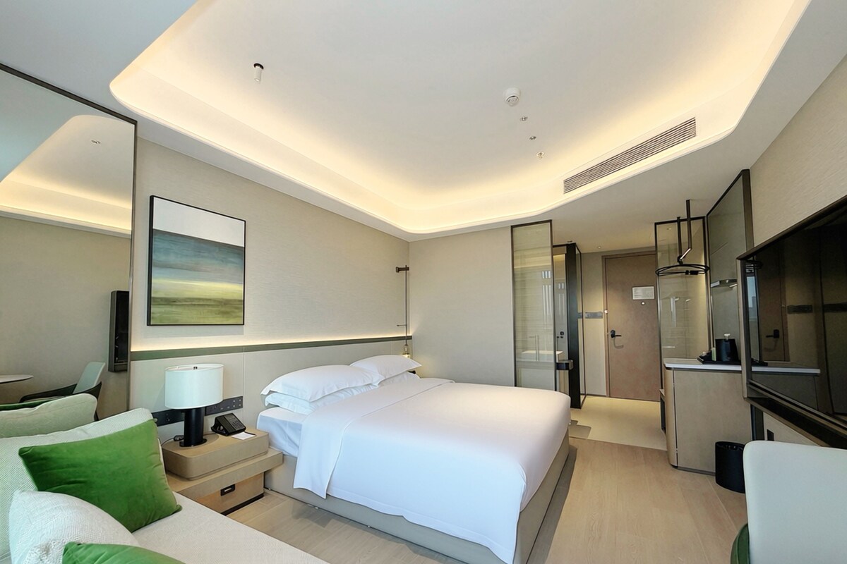 A modern hotel room with a bed, bedside table, lamp, and a painting on the wall.