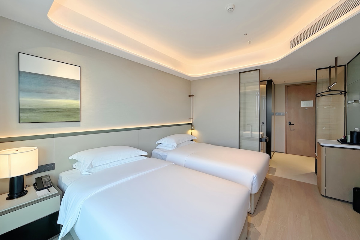 A hotel room with two beds, a bedside table with a lamp, a phone, and a painting on the wall.