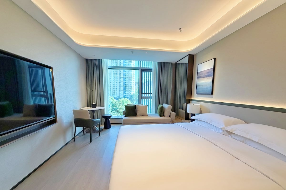 A modern hotel room with a large bed, a seating area with a table, and a window with curtains.