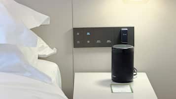 A hotel room with a bed, a bedside table with a speaker, and a control panel with buttons for room settings.