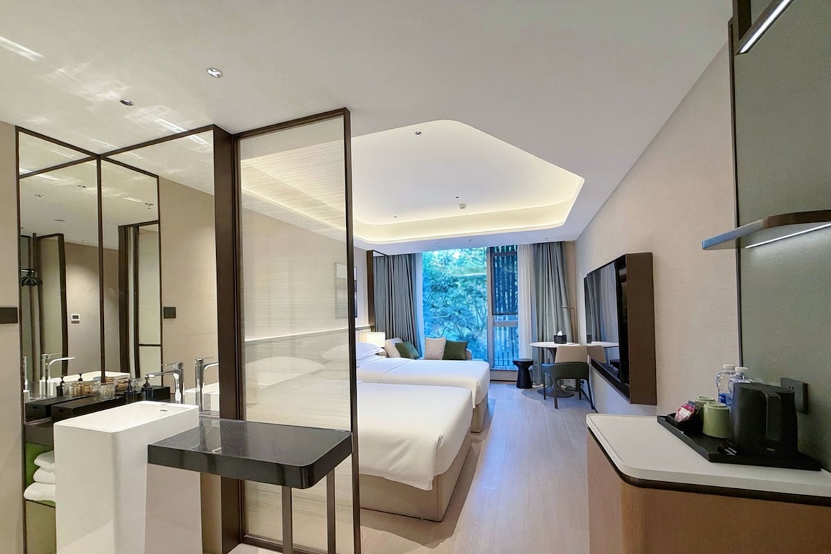 A modern hotel room with a large bed, a separate sitting area, a bathroom with a vanity, and a large mirror.