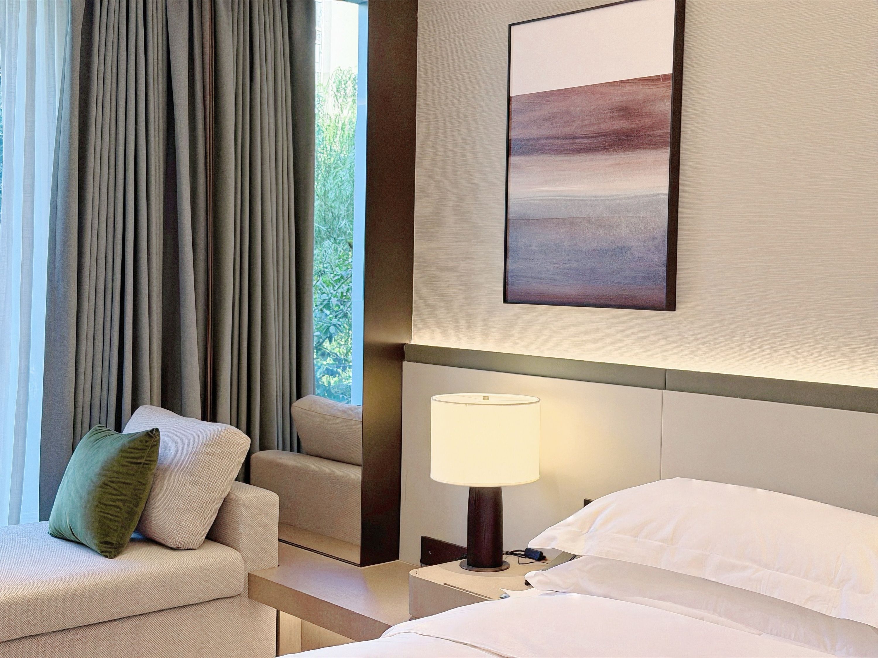 A hotel room with a bed, a nightstand with a lamp, a sofa with cushions, and a window with curtains.